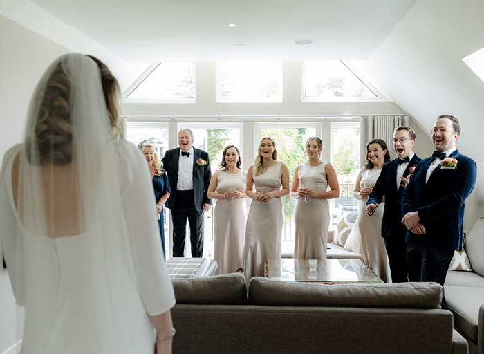 a line of people standing in front of a window wearing cream bridesmaid dresses and smart black dinner suits looking surprised as a bride shows off her dress