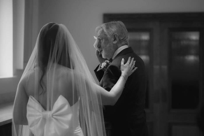 A bride wearing a veil and a large bow on the back of her dress faces away from the camera and comforts her father as he wipes his eyes with a tissue