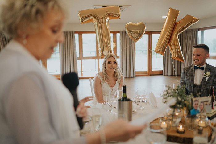 A Lady Stands Holding A Microphone In Foreground As Bride And Groom Sit At Round Table And Watch On With Gold T And K Balloons In Background