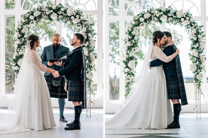 Bride and groom hold hands and hug during wedding ceremony with white floral arch behind them