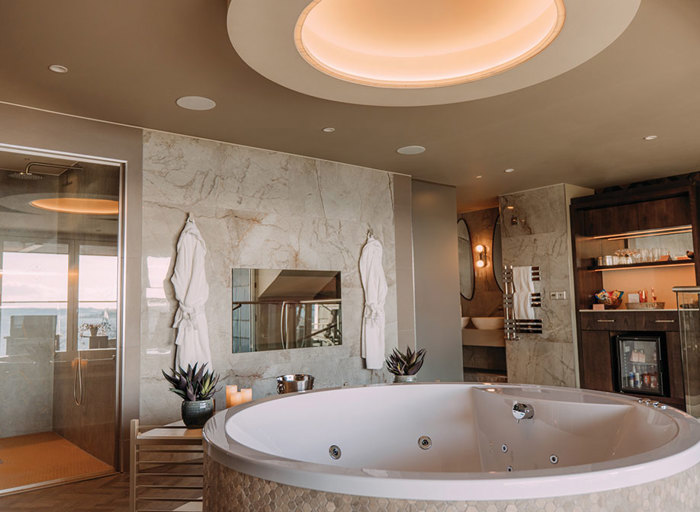 A large bathroom with a spa bath in the foreground, a shower room next to a wall with a TV and two dressing gowns hanging up on the left and a mini bar on the wall to the right