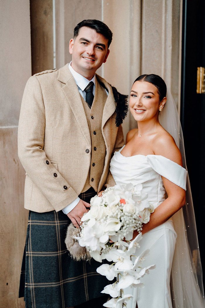 bride in strapless wedding dress with off the shoulder straps holds trailing bouquet while standing beside tall groom in beige and dark green kilt outfit