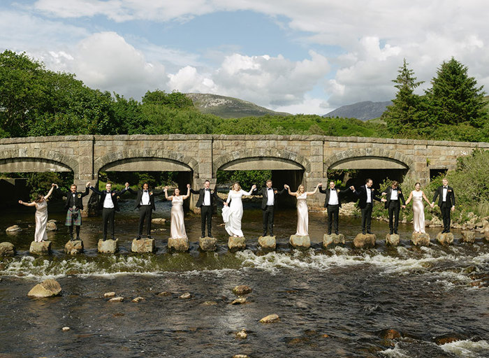 a line of people wearing smart black dinner suits and bow ties and long cream dresses standing on stepping stones across a river. They are holding raised hands in the air and there is an arched stone bridge, trees and hills in background