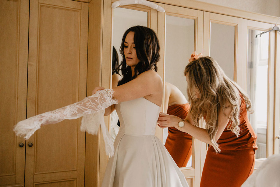 A Person Putting A Wedding Dress On As A Person In An Orange Dress Fastens Its Buttons