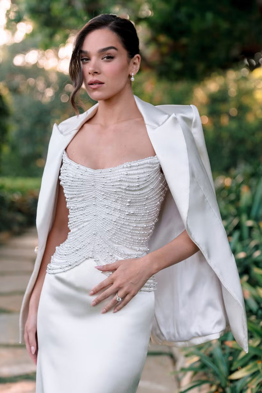 brunette woman wears white blazer over a wedding dress with pearl covered bodice silky skirt standing in front of green planted background