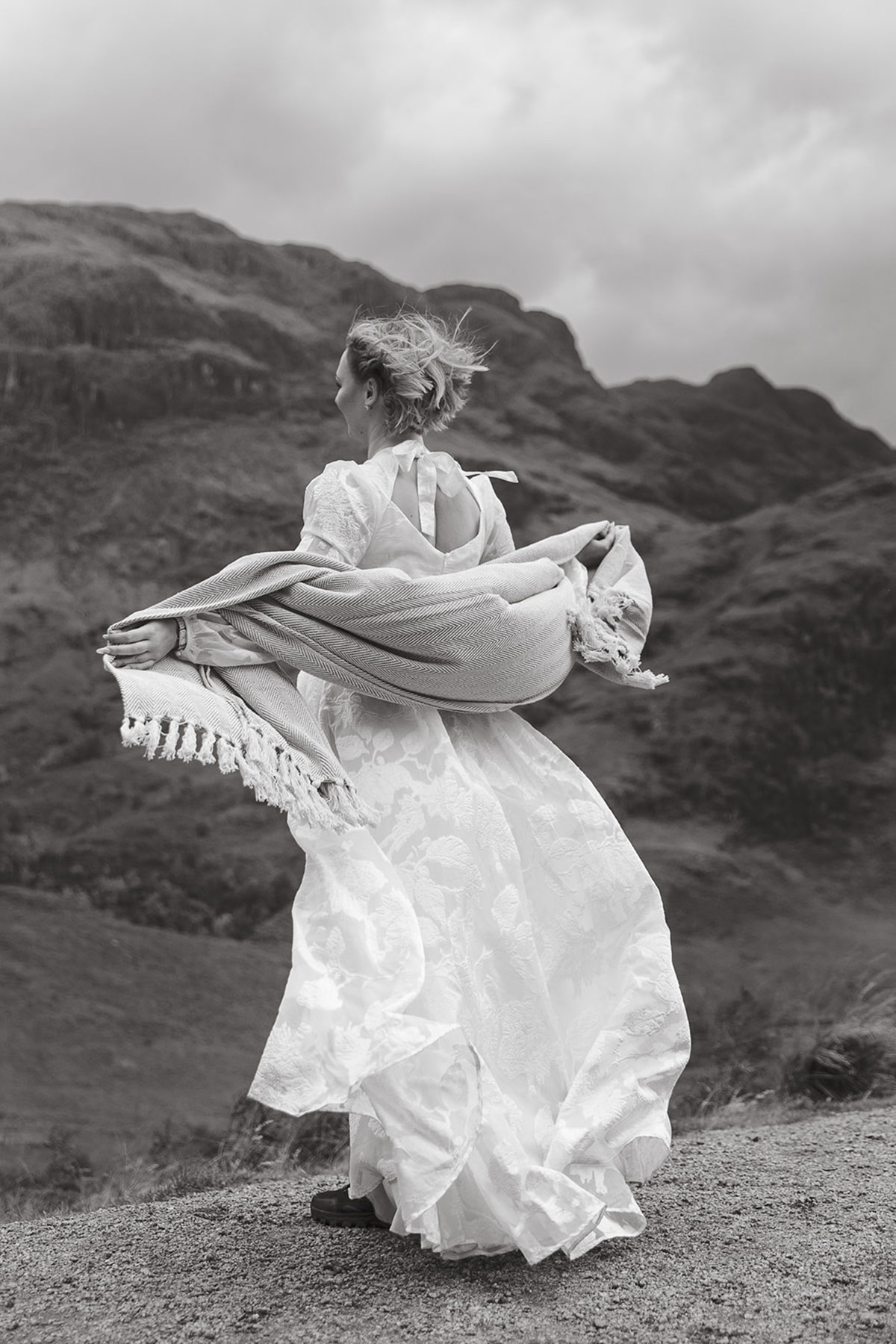 bride with shoulder length hair and floral embellished wedding dress holds a shawl across her arms, extending them outwards, facing the wind and view of nearby hills