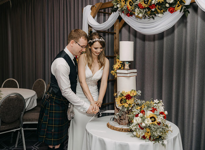 A bride and groom cutting into a three tier cake holding the knife together with flowers around the table 