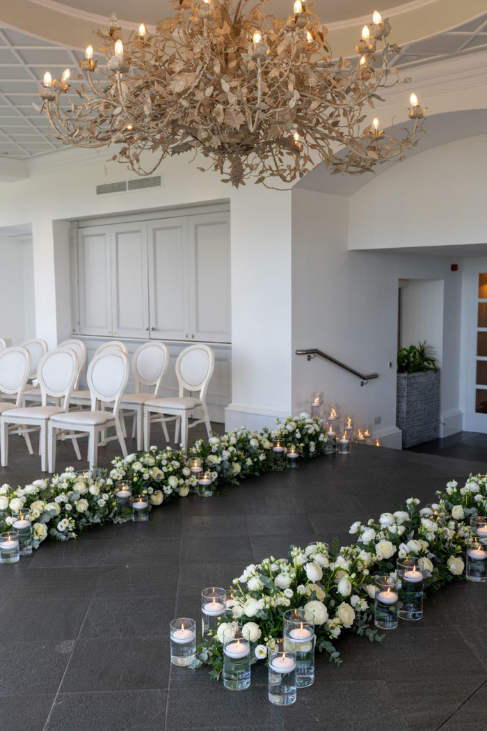 white flower decorations and floating candles in glass holders on a grey tiled floor. Rows of white chairs in background.