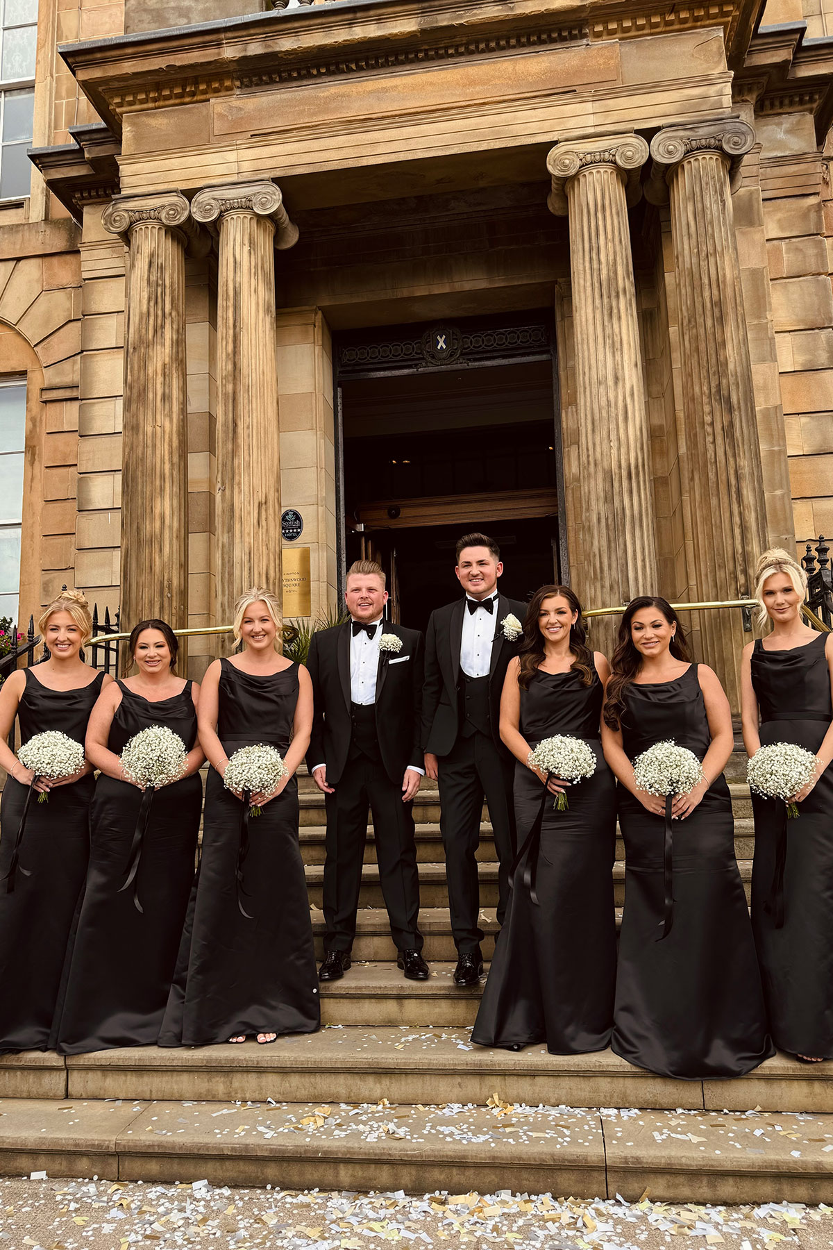 two grooms stand with bridesmaids wearing black dresses on steps at kimpton blythswood square glasgow