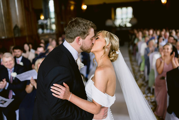 A bride and groom kissing as rows of wedding guests behind them clap.