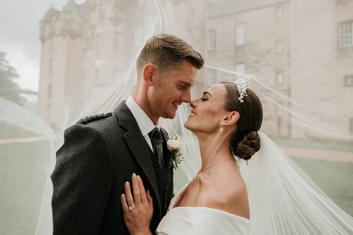 A close up photo of a bride and groom underneath the bride's veil with a castle in the background  