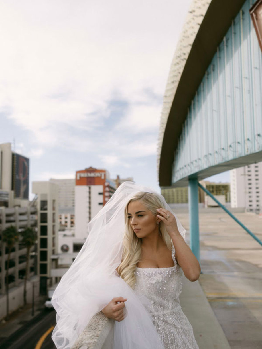 bride holds her long veil over one arm as she runs her other hand through her hair, posing along the side of building vantage point