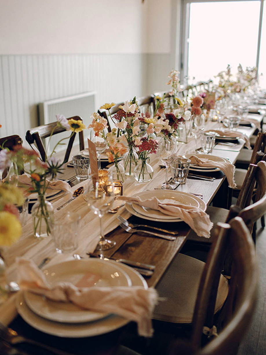 long wooden dining table with chairs lined underneath, white table runner and lots of bud vases down the middle