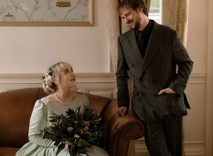 a smiling bride seated on a brown leather couch. She is looking upwards towards a groom wearing a dark tweed suit standing next to her