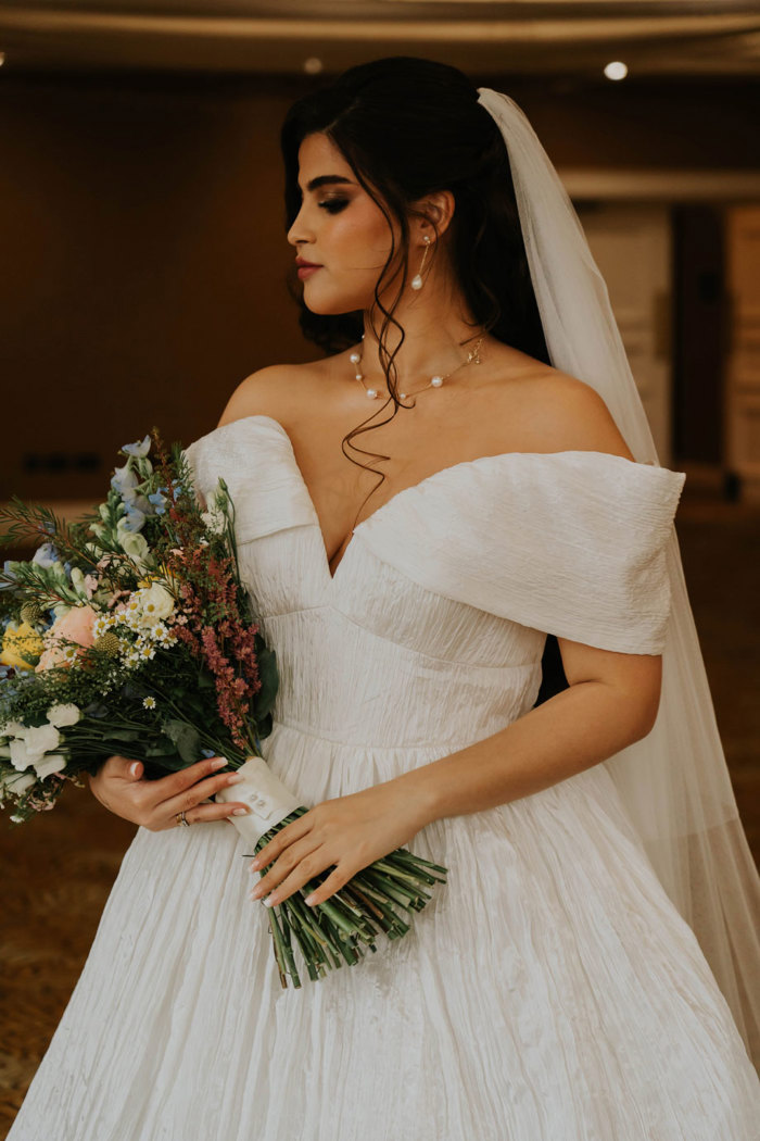 A bride wearing an off-the-shoulder wedding dress and a veil holding a bouquet of wildflowers 