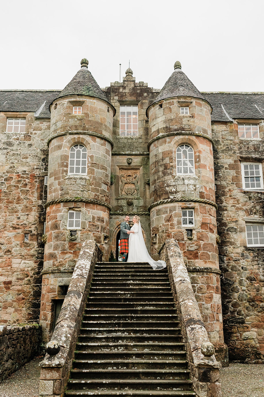 bride in white wedding dress and long veil and groom in red and green tartan kilt stand at the top of a large staircase leading up to a large stone brick castle