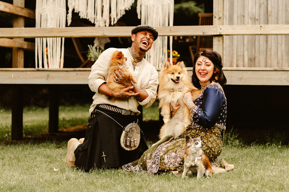 A man in a kilt and a woman in a sari sit on grass cuddling a dog each while another small dog sits next to the woman