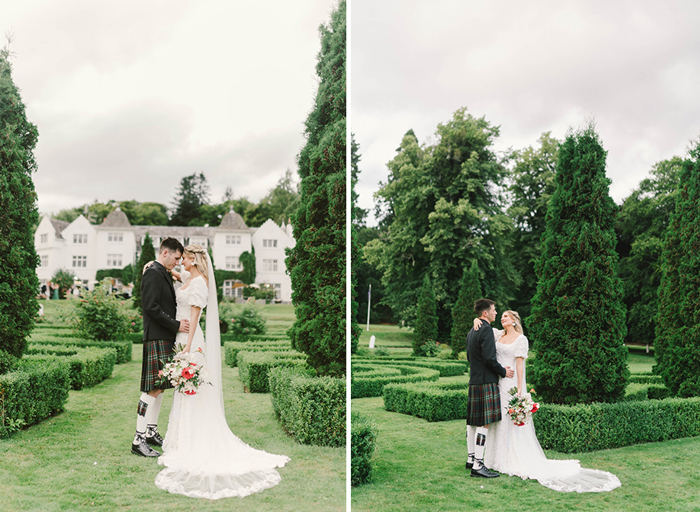 A bride and groom stand outside in a garden with small hedges and fir trees