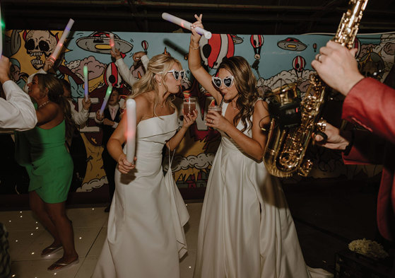 two brides wearing love heart sunglasses dancing with glow sticks on a light-up dance floor. They are both drinking cocktails through a straw. There is a saxophone in the foreground at the right hand side of the image