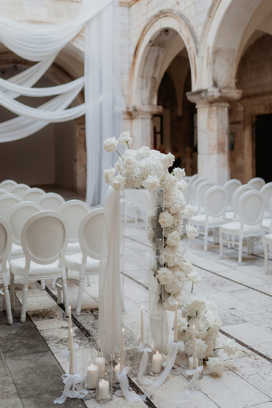 a tall clear perspex stand in a stone courtyard dressed with white roses and glass holders tied with bows filled with candles. There are rows of white chairs in the background