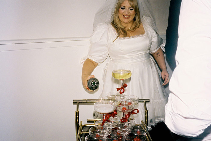 blonde bride pours champagne into champagne tower of coupe glasses with red bows around the stems, all placed on a cocktail trolley