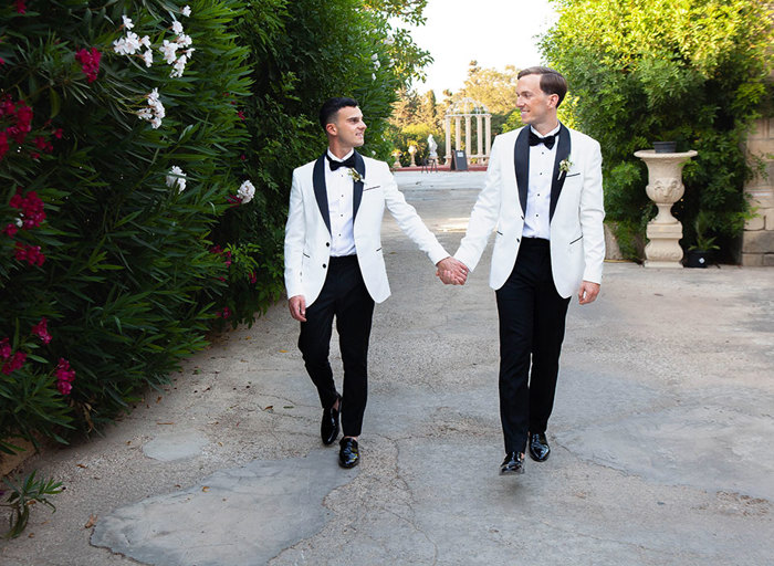 two grooms wearing white suit jackets and bow ties walking along a stone pathway with green hedges blooming with flowers either side