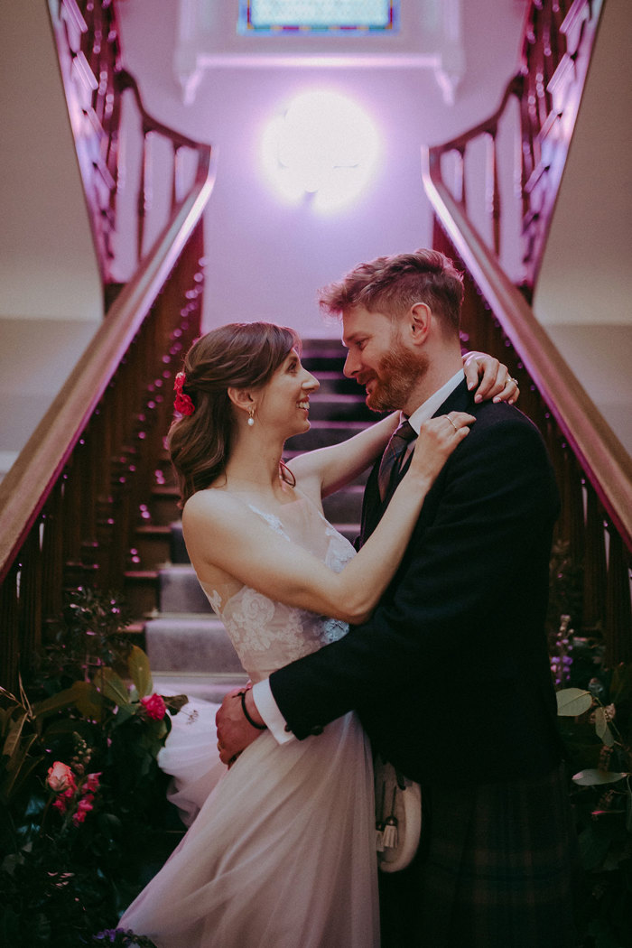 a smiling bride and groom hugging at the bottom of a wooden staircase