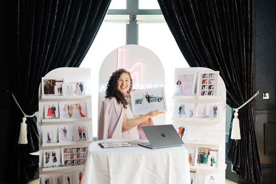 a brunette woman sits behind a table and macbook, smiling and surrounded by a display of printed out portraits