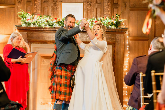 bride in white wedding dress and veil and groom in red and green tartan kilt outfit excitedly hold arms up and out in front of each other with excited looks on their faces