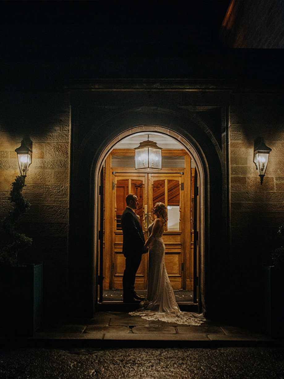 A bride in a long dress and a groom in a suit stand inside a lit doorway at night