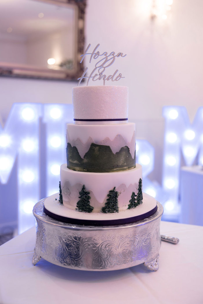 a wedding cake featuring green trees and mountains decoration with a silver script cake topper that reads 'Hozza and Hendo'