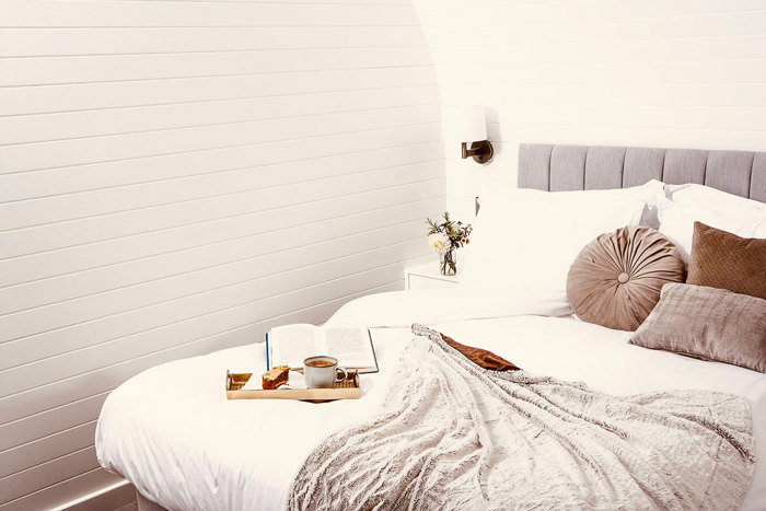 a white sheeted bed with brown pillows, a grey blanket, an open book and a tray with coffee and cake sat on top, situated in the corner of a white panelled room