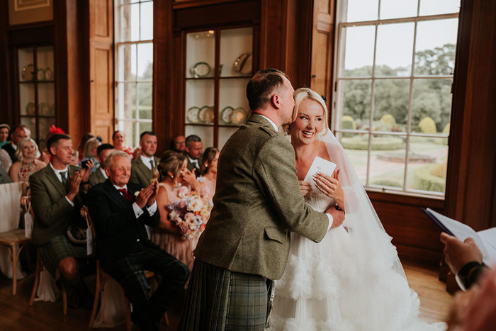 A man in a kilt kisses a bride on the cheek during a wedding ceremony