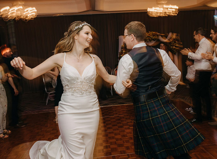 A bride in a white dress and a groom in a kilt link arms and spin around a dance floor