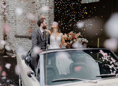 bride and groom sit in cream coloured convertible car wearing heart shaped sunglasses and surrounded by colourful falling confetti