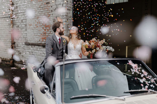 bride and groom sit in cream coloured convertible car wearing heart shaped sunglasses and surrounded by colourful falling confetti