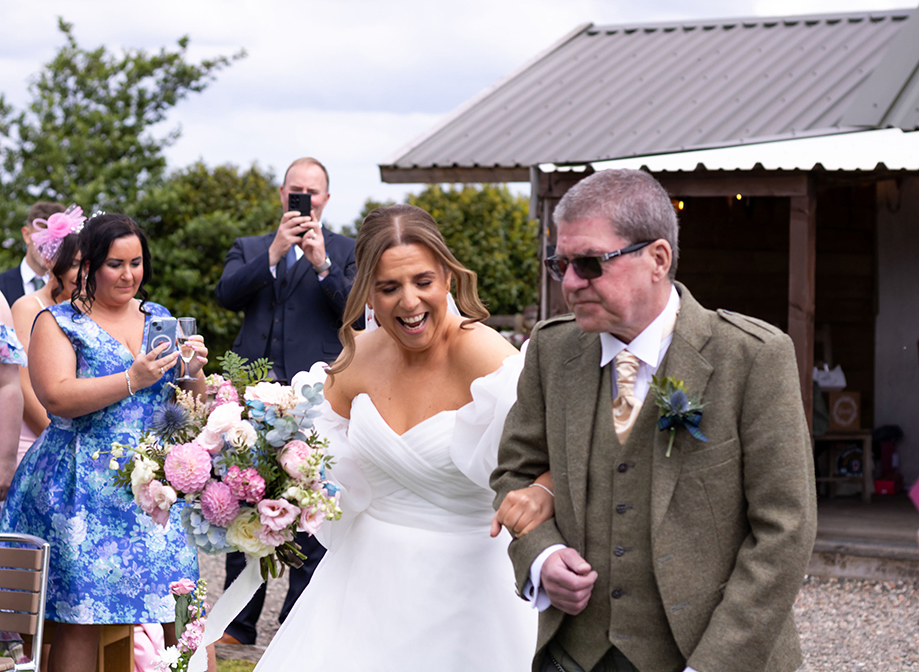 bride laughing and carrying flowers whilst holding onto older man in kilt