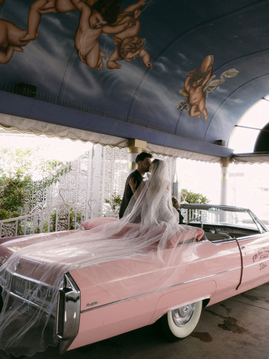 bride and groom sit atop the top of stationary pink convertible car with bride's long veil trailing along the back of car