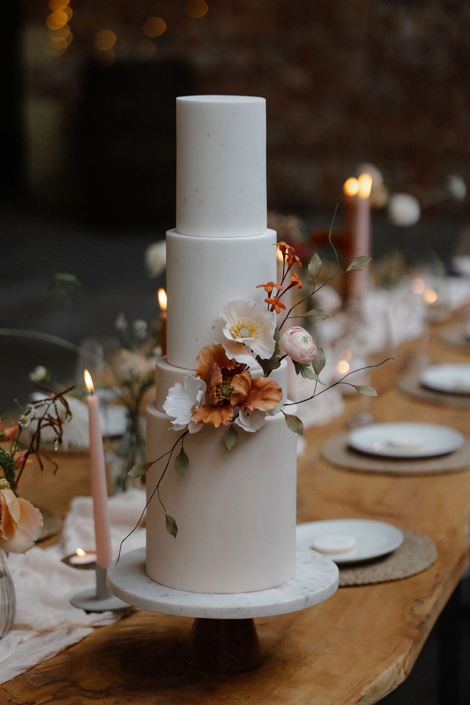 a smooth white four-tiered wedding cake decorated with a few realistic sugar flowers sits on a marble cakestand on a long wooden dining table