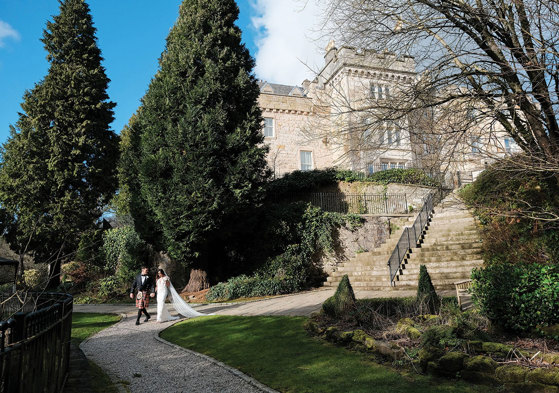 couple walk hand in hand during wedding with exterior of crossbasket castle in the background
