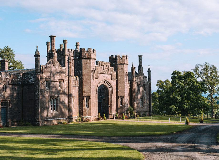 A castle with a green lawn in front of it and sunlight hitting the facade through trees