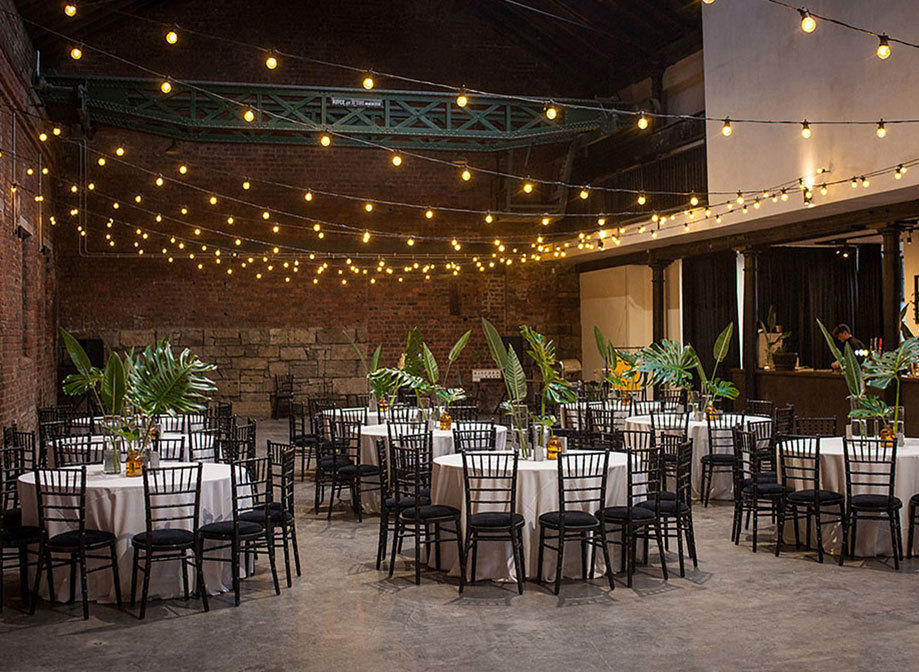 A room with brickwork walls and a selection of round tables with white table cloths and black wooden chairs with plants on the centre of every table