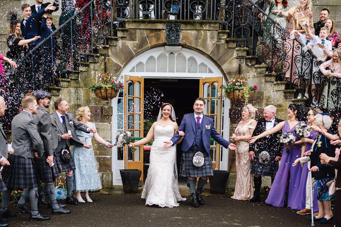 a bride and groom walking through a confetti shower below a stone staircase with wrought iron balustrade.