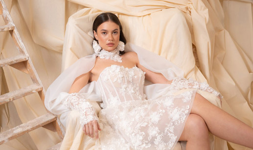bride with dark brown hair wears strapless floral applique mini wedding dress and long matching sleeves as she sits in a chair covered by a dark cream sheet doubling as the backdrop