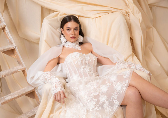 bride with dark brown hair wears strapless floral applique mini wedding dress and long matching sleeves as she sits in a chair covered by a dark cream sheet doubling as the backdrop