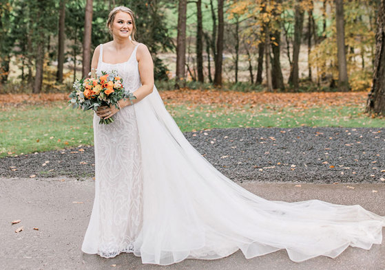 bride holding bouquet stands outside in sequinned wedding dress with long train and veil laid out to the side 