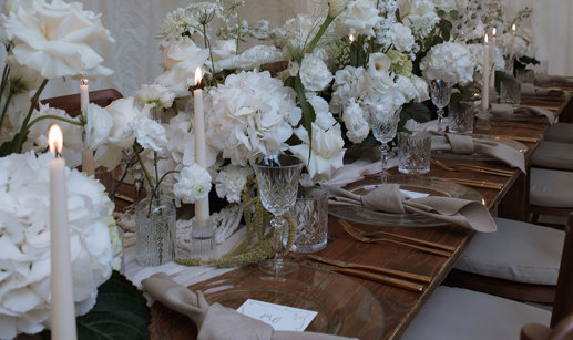 long wooden table set with clear charger plates, linen napkins, and a long middle section filled with white flowers and crystal glassware