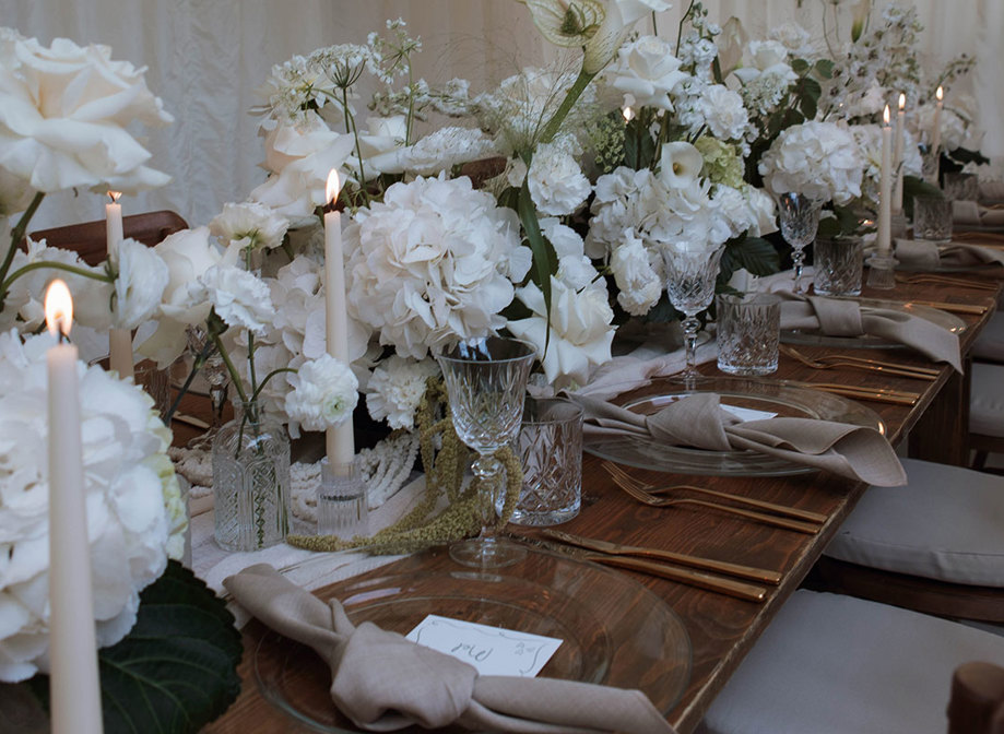 long wooden table set with clear charger plates, linen napkins, and a long middle section filled with white flowers and crystal glassware