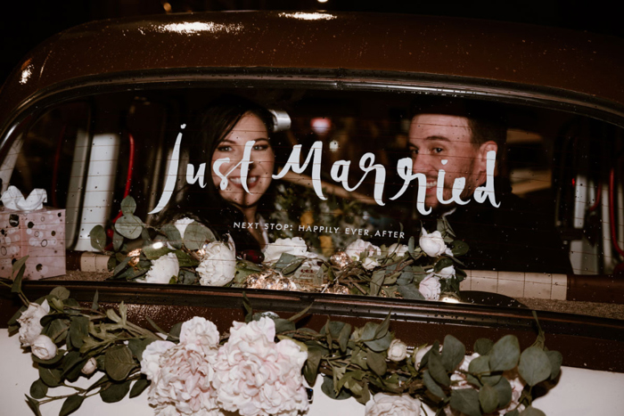 The bride and groom smile out the back window of their white wedding taxi that has Just Married written on it