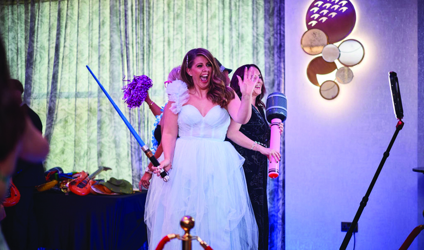 a bride and three other people stand on a 360 photobooth podium holding an array of props as the camera swings around